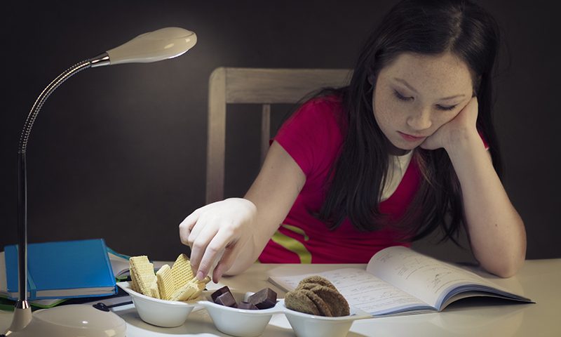 Young girl eating candy while learning in the night (focus is set on hand taking candy)